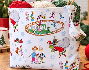 May include: A white fabric storage bag featuring a colorful print of characters from How the Grinch Stole Christmas, including the Grinch, Cindy Lou Who, and a train on tracks, accented with blue snowflakes and two wooden knitting needles.