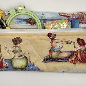 May include: A rectangular quilted fabric zipper pouch featuring a beach scene print with figures in swimwear. The pouch contains a light green embroidery hoop and yellow embroidery floss. A silver starfish charm is attached to the zipper pull.
