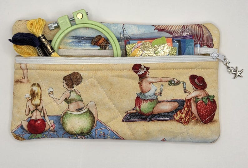 May include: A rectangular quilted fabric zipper pouch featuring a beach scene print with figures in swimwear. The pouch contains a light green embroidery hoop and yellow embroidery floss. A silver starfish charm is attached to the zipper pull.