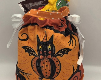 May include: A fabric drawstring bag featuring an orange background with a black cat and pumpkin graphic. The bag has a top handle with white polka dots and white ribbon ties on the sides. The bag is filled with yellow and brown candy wrappers.