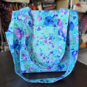 Puede incluir: Un bolso tote acolchado con un vibrante estampado floral en tonos turquesa, azul claro y morado. El bolso incluye dos correas largas para el hombro y un bolsillo exterior centrado, todo cubierto con un diseño repetitivo de flores pequeñas.