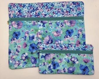 May include: Two rectangular fabric pouches featuring a quilted floral pattern in shades of blue, purple, and teal. Each pouch has a teal zipper closure at the top, with the larger pouch positioned behind the smaller one on a plain background.