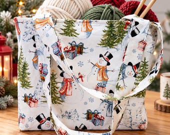 May include: A white fabric tote bag featuring a repeating pattern of snowmen wearing hats and scarves, green pine trees, and colorful gift boxes. The bag is filled with several balls of yarn in cream, red, and green, along with two wooden knitting needles.