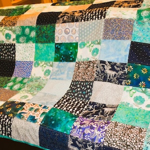 May include: A patchwork quilt with a variety of green, blue, and neutral-toned fabrics. The quilt features a mix of patterns, including floral, geometric, and abstract designs. The edges are finished with a teal border. The quilt is draped over a dark surface.