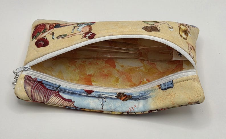 May include: A rectangular fabric zipper pouch featuring a beige exterior with a repeating illustration of figures in red and blue clothing. The interior is lined with a light orange and yellow patterned fabric, and a white zipper runs across the top.