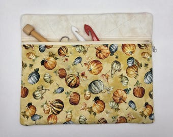 May include: A rectangular quilted fabric pouch featuring a repeating pattern of orange, green, and blue pumpkins on a yellow background. The pouch has a top zipper closure and contains a wooden tool, a white hook, and a red hook inside.