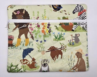 May include: A quilted zippered pouch in light green featuring a colorful garden print with a bison, zebra, mouse, dachshund, squirrel, and goat. The fabric displays various plants and flowers, and the text Cross Stitch Bag 1 is printed in white.