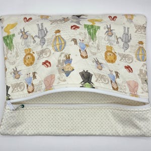 Puede incluir: Un estuche rectangular acolchado con cremallera hecho de tela color crema, estampado con varios personajes e iconos del Mago de Oz, incluyendo a Dorothy, el Hombre de Hojalata, el Le&oacute;n, globos aerost&aacute;ticos y zapatos rojos, con cremallera blanca.