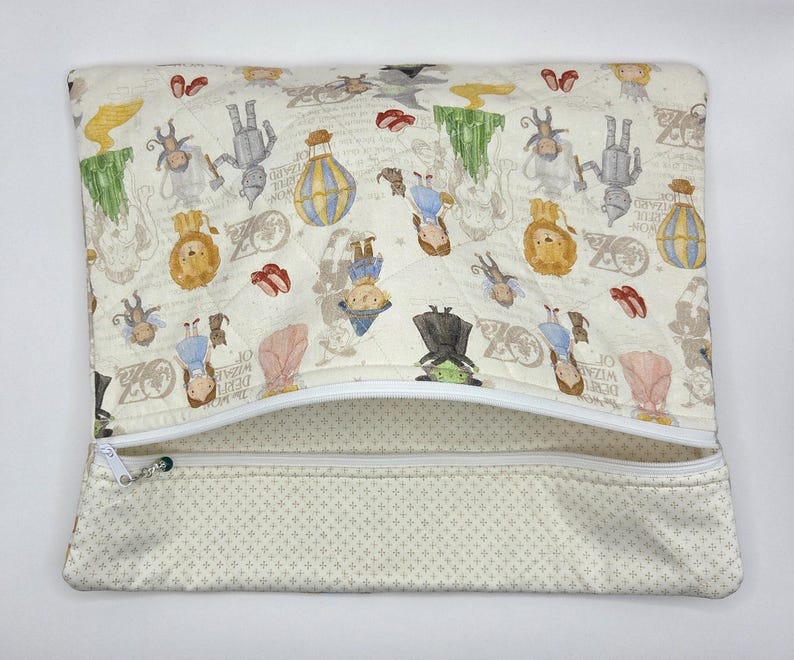 Puede incluir: Un estuche rectangular acolchado con cremallera hecho de tela color crema, estampado con varios personajes e iconos del Mago de Oz, incluyendo a Dorothy, el Hombre de Hojalata, el Le&oacute;n, globos aerost&aacute;ticos y zapatos rojos, con cremallera blanca.