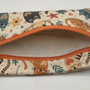May include: A rectangular fabric zipper pouch featuring a cream-colored background with a repeating pattern of stylized cats, flowers, and leaves in shades of orange, gray, and teal. The pouch has an orange zipper and a small silver flower charm.