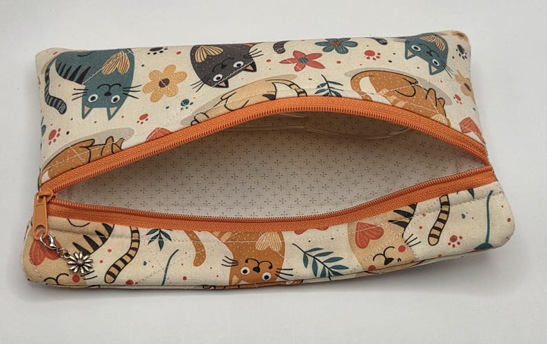May include: A rectangular fabric zipper pouch featuring a cream-colored background with a repeating pattern of stylized cats, flowers, and leaves in shades of orange, gray, and teal. The pouch has an orange zipper and a small silver flower charm.