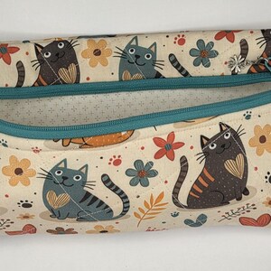 May include: A rectangular fabric zipper pouch featuring a cream background with a repeating pattern of gray and brown cats, orange and blue flowers, and small paw prints. The pouch has a teal zipper closure and a small silver flower charm.