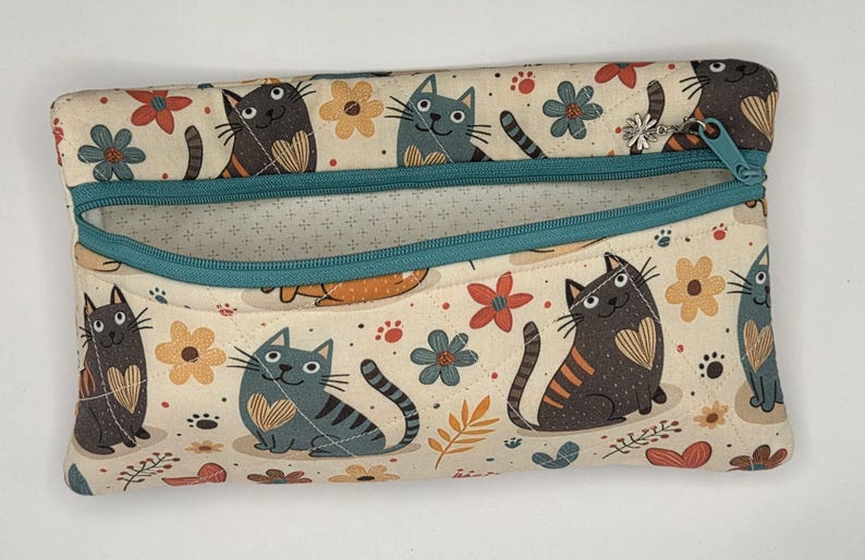 May include: A rectangular fabric zipper pouch featuring a cream background with a repeating pattern of gray and brown cats, orange and blue flowers, and small paw prints. The pouch has a teal zipper closure and a small silver flower charm.