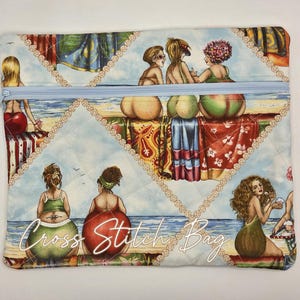 May include: A rectangular quilted fabric pouch with a light blue zipper closure. The surface features a colorful beach scene print with multiple figures sitting on towels by the ocean. The text Cross Stitch Bag is printed in white script.