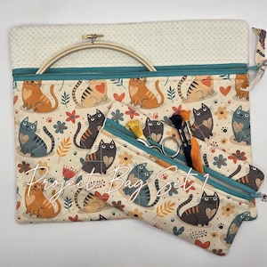 May include: A project bag set featuring a cream-colored fabric with a repeating pattern of orange and gray cats, hearts, and flowers. The bags have teal zippers and include a wooden embroidery hoop, metal scissors, and embroidery floss.