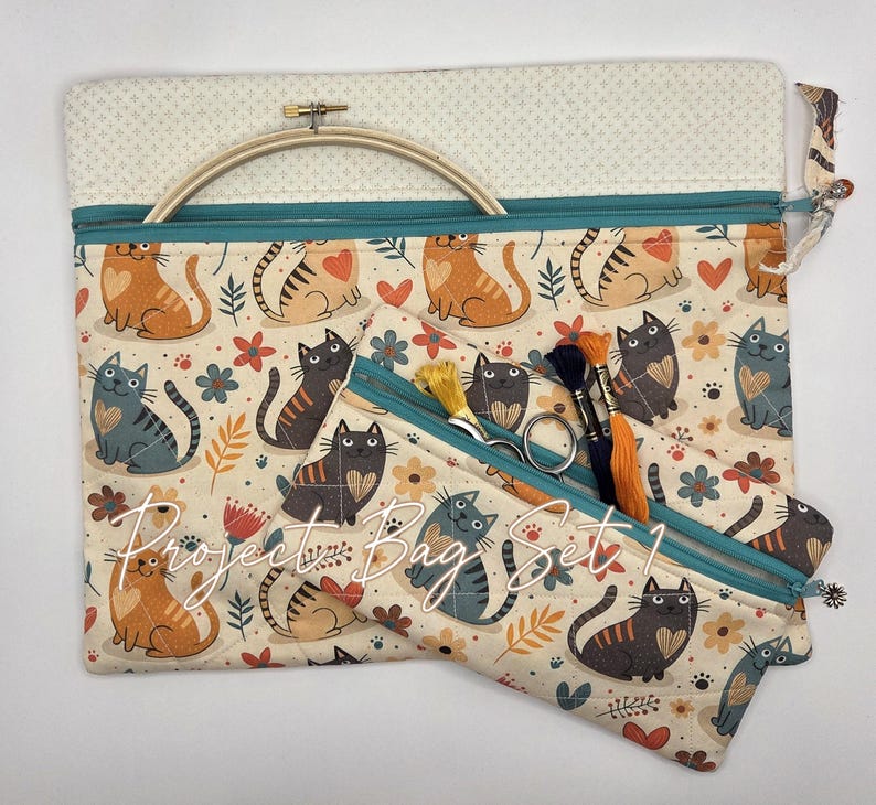 May include: A project bag set featuring a cream-colored fabric with a repeating pattern of orange and gray cats, hearts, and flowers. The bags have teal zippers and include a wooden embroidery hoop, metal scissors, and embroidery floss.