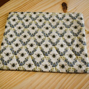 May include: A rectangular, quilted pouch with a repeating pattern of black bees and blue floral accents on a cream background. The pouch has a gold zipper pull with a blue bead. The pouch is lying on a light wood surface.