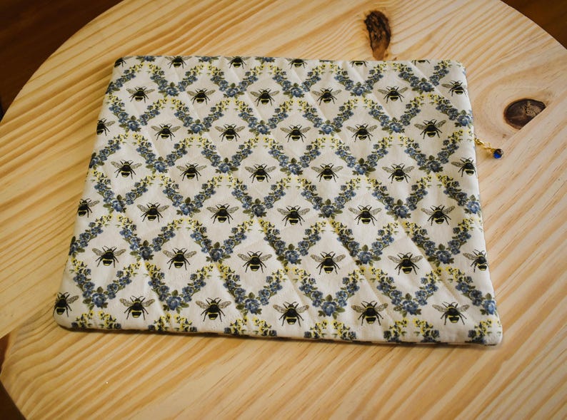 May include: A rectangular, quilted pouch with a repeating pattern of black bees and blue floral accents on a cream background. The pouch has a gold zipper pull with a blue bead. The pouch is lying on a light wood surface.