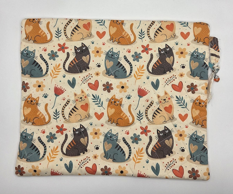 May include: A rectangular fabric pouch featuring a repeating pattern of orange and gray cats with heart-shaped markings on their chests, interspersed with small flowers, leaves, and heart shapes on a cream-colored background with quilted stitching.