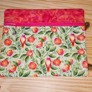 K&ouml;nnte beinhalten: Eine rechteckige, gesteppte Stoffh&uuml;lle mit einem rosa Rei&szlig;verschluss. Der obere Teil ist ein leuchtendes Pink mit einem Rautenmuster. Der Hauptteil zeigt ein Blumenmuster mit roten V&ouml;geln, rosa und orangefarbenen Rosen und gr&uuml;nen Bl&auml;ttern.