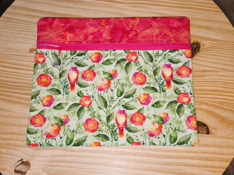 K&ouml;nnte beinhalten: Eine rechteckige, gesteppte Stoffh&uuml;lle mit einem rosa Rei&szlig;verschluss. Der obere Teil ist ein leuchtendes Pink mit einem Rautenmuster. Der Hauptteil zeigt ein Blumenmuster mit roten V&ouml;geln, rosa und orangefarbenen Rosen und gr&uuml;nen Bl&auml;ttern.