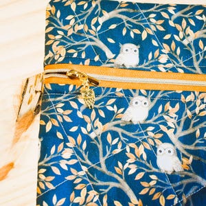 K&ouml;nnte beinhalten: Eine gesteppte, marineblaue Stofftasche mit goldenem Rei&szlig;verschluss und Eulenanh&auml;nger. Der Stoff zeigt ein sich wiederholendes Muster aus wei&szlig;en Eulen auf &Auml;sten mit goldenen Bl&auml;ttern. Die Tasche hat eine quadratische Form.