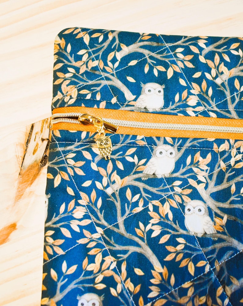 K&ouml;nnte beinhalten: Eine gesteppte, marineblaue Stofftasche mit goldenem Rei&szlig;verschluss und Eulenanh&auml;nger. Der Stoff zeigt ein sich wiederholendes Muster aus wei&szlig;en Eulen auf &Auml;sten mit goldenen Bl&auml;ttern. Die Tasche hat eine quadratische Form.
