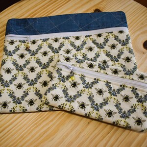 May include: Two zippered pouches. The larger pouch has a denim blue top and a white zipper. The fabric features a pattern of black bees and blue floral accents on a cream background. The smaller pouch has the same pattern.