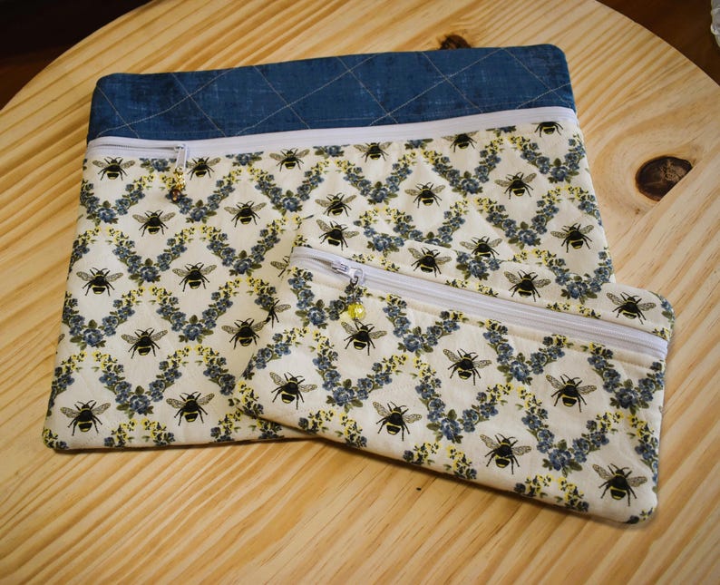 May include: Two zippered pouches. The larger pouch has a denim blue top and a white zipper. The fabric features a pattern of black bees and blue floral accents on a cream background. The smaller pouch has the same pattern.