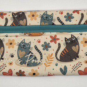 May include: A rectangular fabric pouch featuring a repeating pattern of cats and flowers on a cream background. The pouch has a teal zipper closure with a small silver flower charm attached. The text "Notions Pouch 1" is printed on the front.