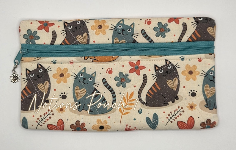 May include: A rectangular fabric pouch featuring a repeating pattern of cats and flowers on a cream background. The pouch has a teal zipper closure with a small silver flower charm attached. The text "Notions Pouch 1" is printed on the front.
