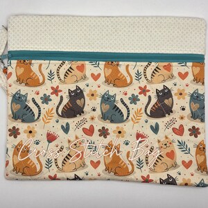 May include: A rectangular quilted pouch featuring a repeating pattern of orange and gray cats, red hearts, and small flowers on a cream background. The bag has a teal zipper closure and a cream-colored top panel with a small geometric print.