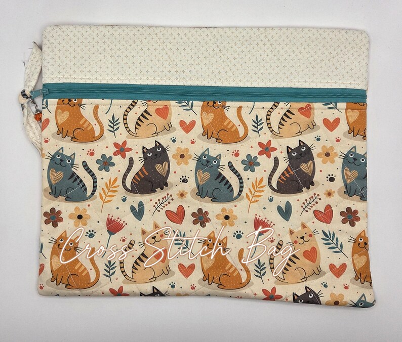 May include: A rectangular quilted pouch featuring a repeating pattern of orange and gray cats, red hearts, and small flowers on a cream background. The bag has a teal zipper closure and a cream-colored top panel with a small geometric print.