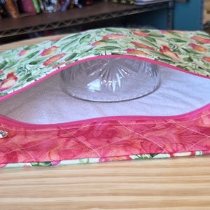 K&ouml;nnte beinhalten: Ein gesteppter Stoffbeutel mit einem Blumenmuster in Rosa-, Gr&uuml;n- und Wei&szlig;t&ouml;nen. Der Beutel hat einen Rei&szlig;verschluss mit einem dekorativen Anh&auml;nger und eine klare Glasschale im Inneren. Der Beutel liegt auf einer Holzoberfl&auml;che.