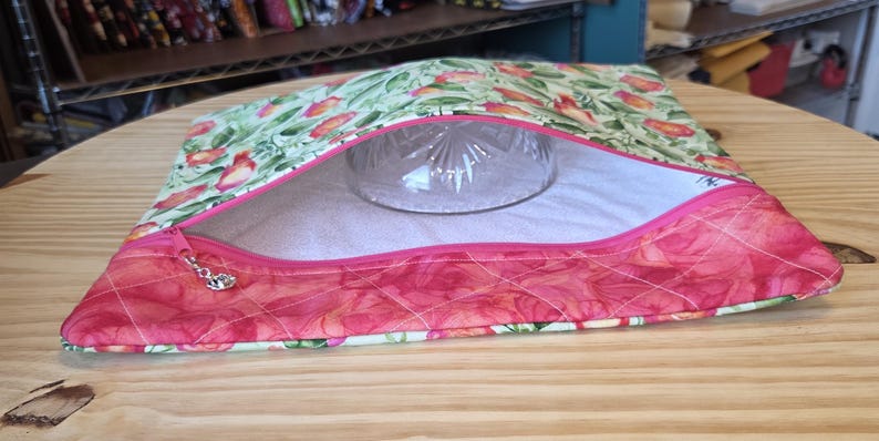 K&ouml;nnte beinhalten: Ein gesteppter Stoffbeutel mit einem Blumenmuster in Rosa-, Gr&uuml;n- und Wei&szlig;t&ouml;nen. Der Beutel hat einen Rei&szlig;verschluss mit einem dekorativen Anh&auml;nger und eine klare Glasschale im Inneren. Der Beutel liegt auf einer Holzoberfl&auml;che.