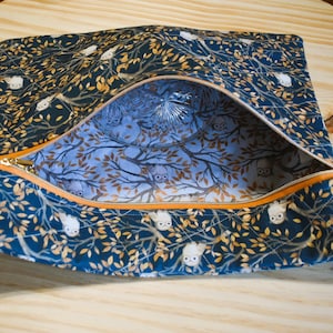 K&ouml;nnte beinhalten: Eine rechteckige, offene Rei&szlig;verschlusstasche. Die dunkelblaue Au&szlig;enseite weist ein Muster aus wei&szlig;en Eulen und goldenen Bl&auml;ttern auf. Der Innenraum ist hellblau mit einem Baum- und Eulenmuster. Der Rei&szlig;verschluss ist orange.
