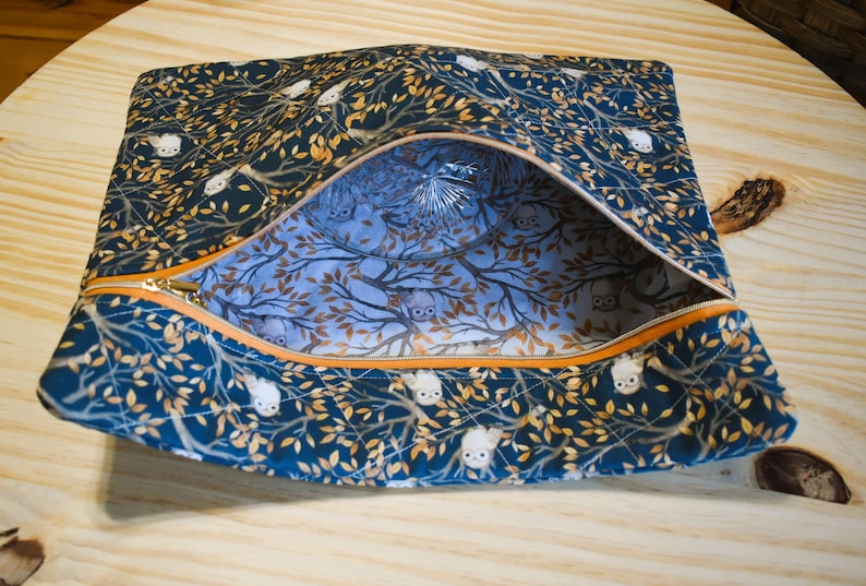 K&ouml;nnte beinhalten: Eine rechteckige, offene Rei&szlig;verschlusstasche. Die dunkelblaue Au&szlig;enseite weist ein Muster aus wei&szlig;en Eulen und goldenen Bl&auml;ttern auf. Der Innenraum ist hellblau mit einem Baum- und Eulenmuster. Der Rei&szlig;verschluss ist orange.