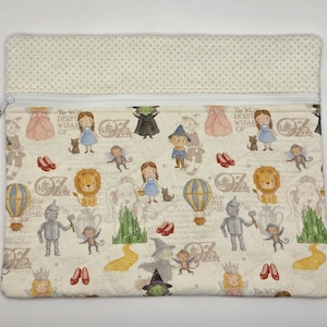 Puede incluir: Un estuche rectangular de tela con cremallera que presenta un patr&oacute;n repetitivo de personajes del Mago de Oz, incluyendo a Dorothy, el Hombre de Hojalata, el Le&oacute;n y la Bruja Malvada, con un panel superior crema y una peque&ntilde;a cuenta verde en el tirador.