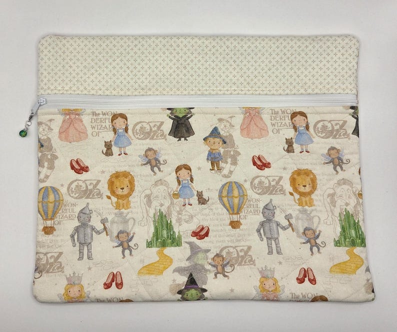 Puede incluir: Un estuche rectangular de tela con cremallera que presenta un patr&oacute;n repetitivo de personajes del Mago de Oz, incluyendo a Dorothy, el Hombre de Hojalata, el Le&oacute;n y la Bruja Malvada, con un panel superior crema y una peque&ntilde;a cuenta verde en el tirador.