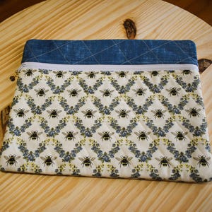 May include: A quilted fabric pouch with a blue denim top and a white zipper. The main body of the pouch features a repeating pattern of black bees and blue floral accents on a white background. The pouch is resting on a wooden surface.