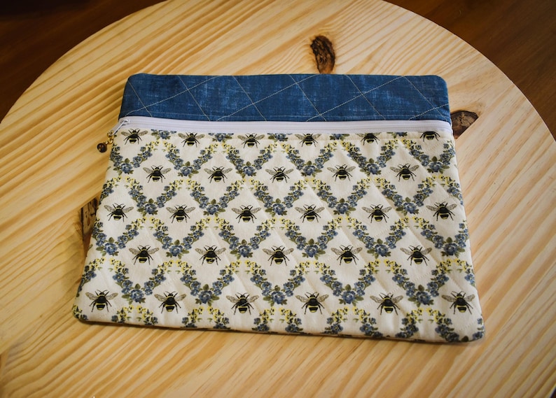 May include: A quilted fabric pouch with a blue denim top and a white zipper. The main body of the pouch features a repeating pattern of black bees and blue floral accents on a white background. The pouch is resting on a wooden surface.