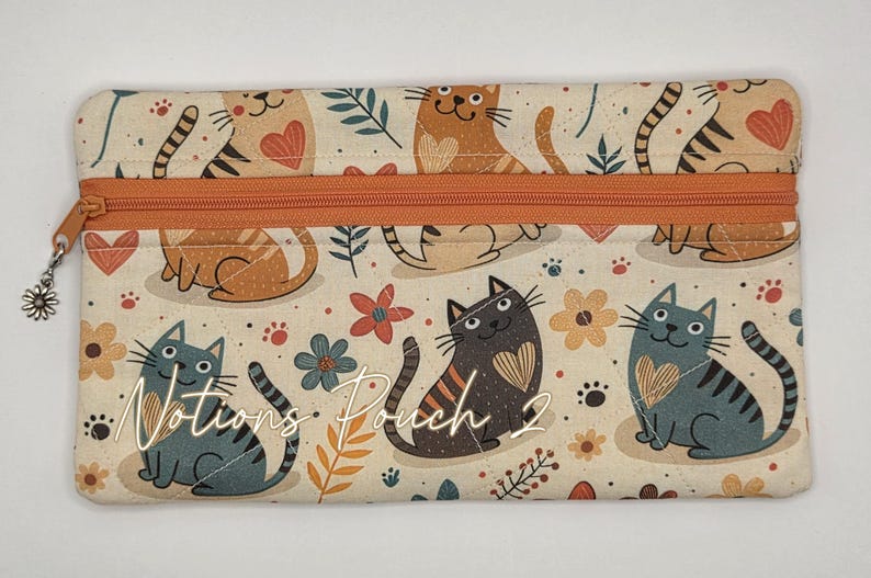 May include: A rectangular fabric notions pouch featuring a cream background with a repeating pattern of cats, hearts, and flowers in shades of orange, gray, and brown. It has an orange zipper closure with a small silver daisy charm and the text Notions Pouch 2.