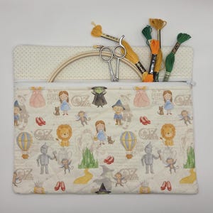 Puede incluir: Un estuche de tela acolchada rectangular con un patr&oacute;n repetitivo de personajes del Mago de Oz, incluyendo a Dorothy, el Hombre de Hojalata, el Le&oacute;n y la Ciudad Esmeralda. El estuche contiene un bastidor de bordado de madera, tijeras e hilos.