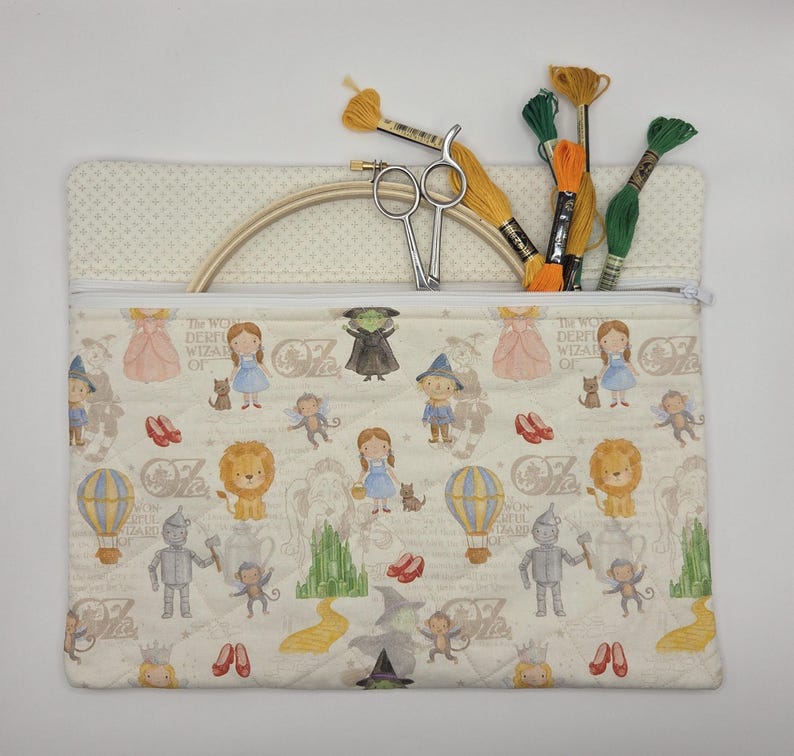 Puede incluir: Un estuche de tela acolchada rectangular con un patr&oacute;n repetitivo de personajes del Mago de Oz, incluyendo a Dorothy, el Hombre de Hojalata, el Le&oacute;n y la Ciudad Esmeralda. El estuche contiene un bastidor de bordado de madera, tijeras e hilos.