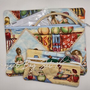 May include: A quilted fabric project bag set featuring two zippered pouches with a beach scene print of people relaxing. The larger bag contains a wooden embroidery hoop and a piece of white fabric. The smaller bag holds metal scissors.