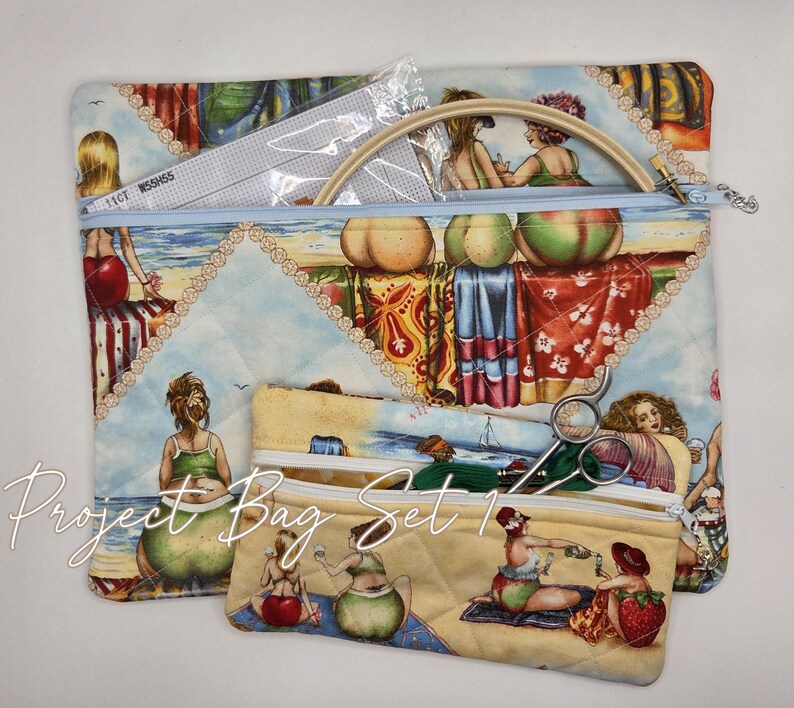 May include: A quilted fabric project bag set featuring two zippered pouches with a beach scene print of people relaxing. The larger bag contains a wooden embroidery hoop and a piece of white fabric. The smaller bag holds metal scissors.