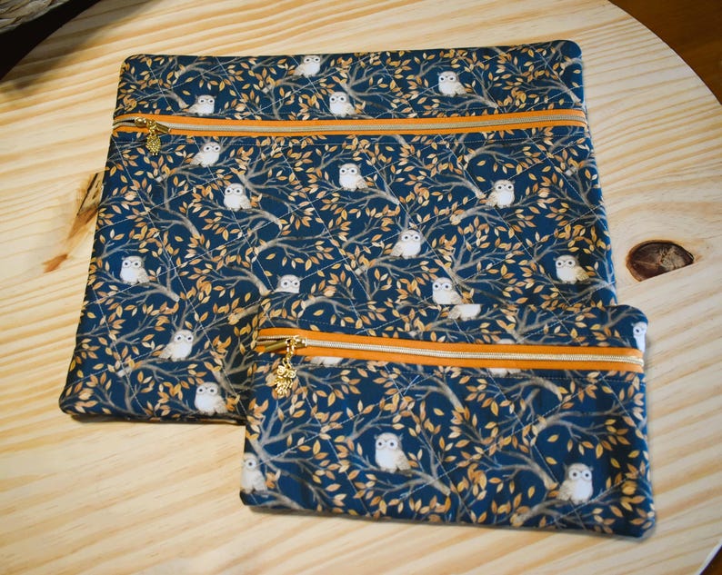 K&ouml;nnte beinhalten: Zwei Rei&szlig;verschlusstaschen mit einem sich wiederholenden Muster aus wei&szlig;en Eulen und goldenen Bl&auml;ttern auf marineblauem Hintergrund. Die Taschen haben orangefarbene Paspeln und goldene Rei&szlig;verschl&uuml;sse.