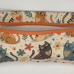 May include: A rectangular fabric zipper pouch featuring a cream background with a repeating pattern of cartoon cats, hearts, and flowers in shades of orange, blue, and dark gray. The pouch has an orange zipper closure with a small silver flower charm.