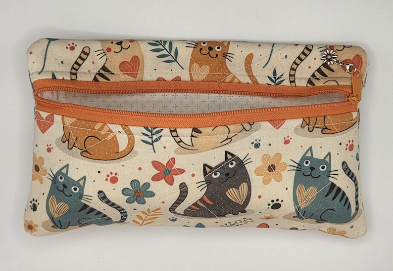 May include: A rectangular fabric zipper pouch featuring a cream background with a repeating pattern of cartoon cats, hearts, and flowers in shades of orange, blue, and dark gray. The pouch has an orange zipper closure with a small silver flower charm.