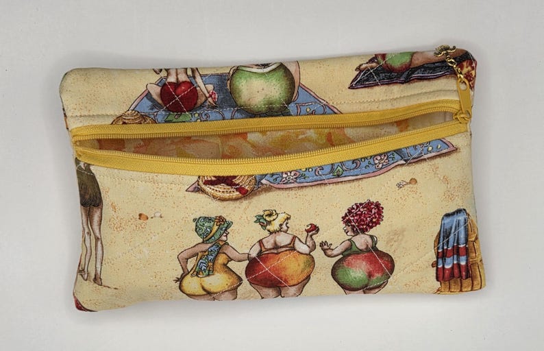 May include: A rectangular quilted fabric pouch featuring a beige background with a repeating print of cartoon figures in swimwear on a beach. The pouch has a yellow zipper closure along the top edge and a small gold-toned metal charm on the zipper pull.